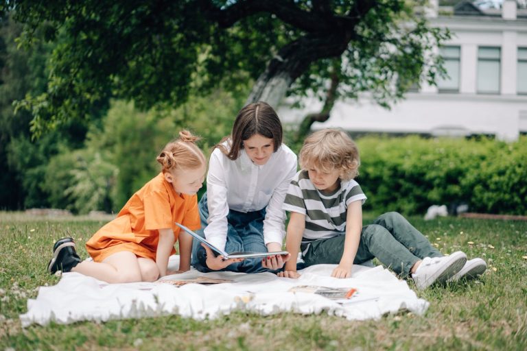 Three kids reading a book on a picnic blanket in a park under a tree.
