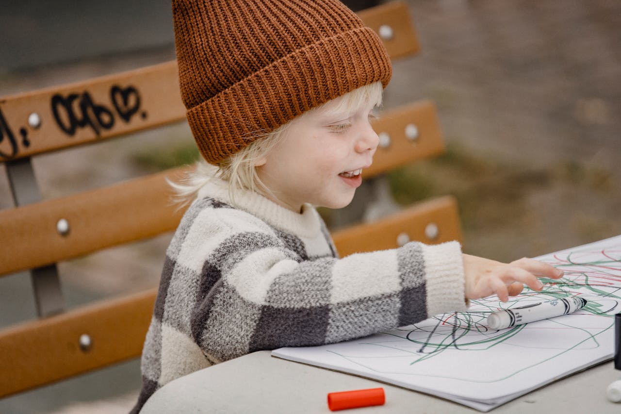 A cheerful child in a beanie drawing with markers at an outdoor park.